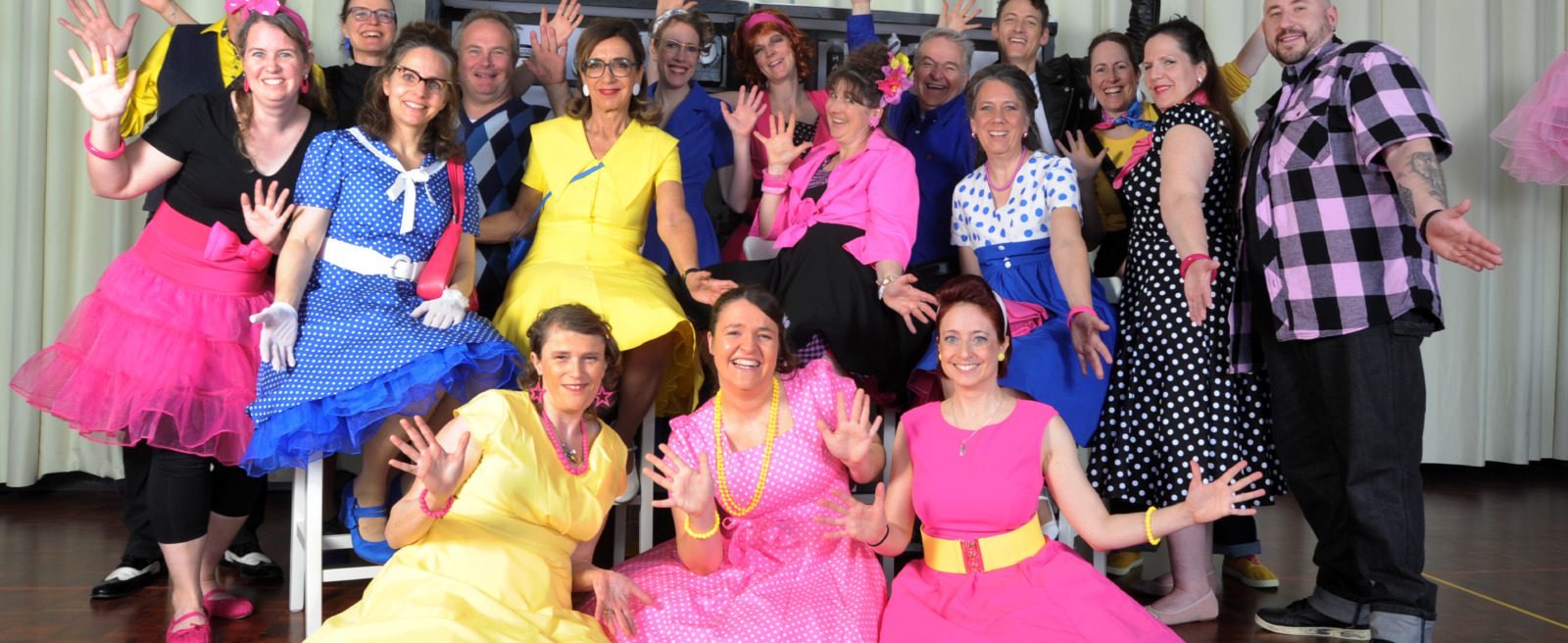 A large group of people in bright 1950s-style dresses posing with hands raised in joy on a wooden floor in front of a light curtain.