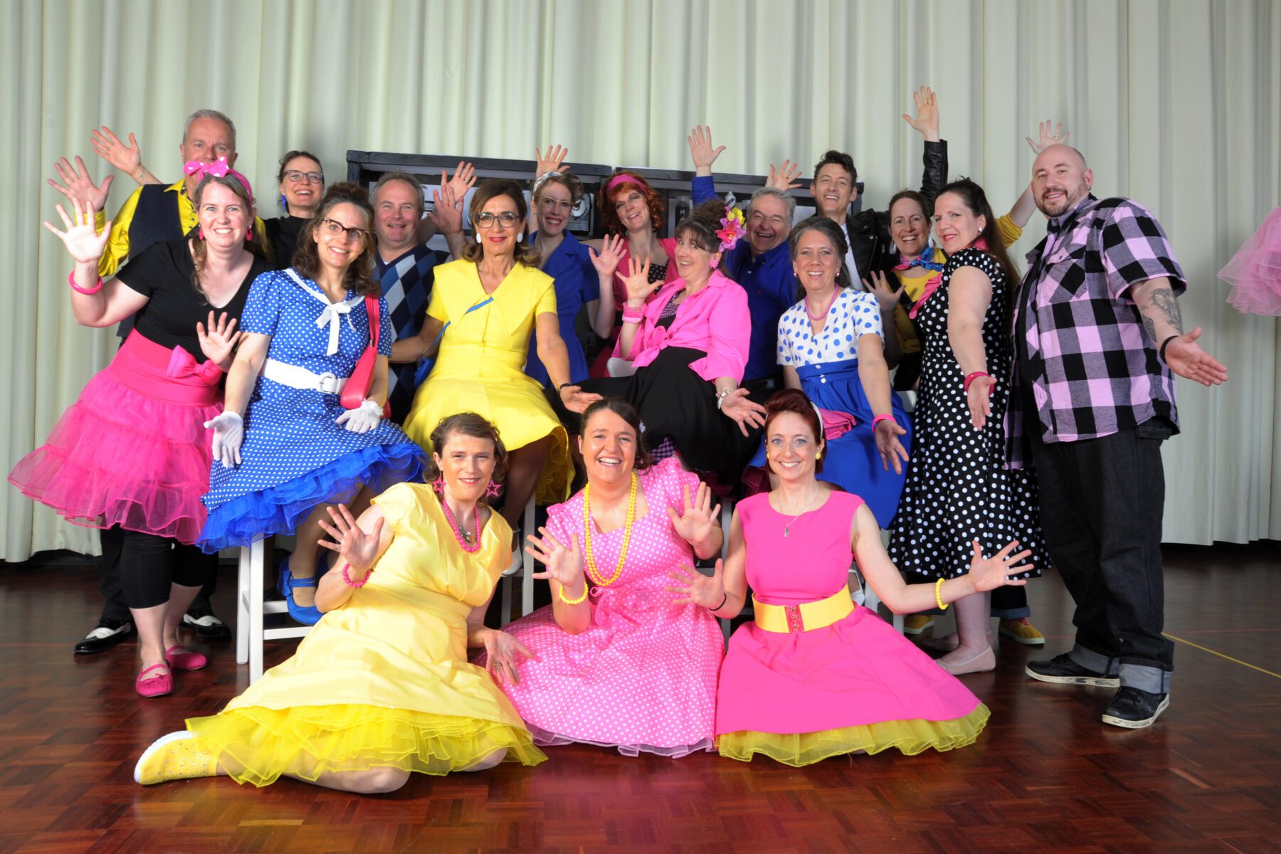 A large group of people in bright 1950s-style dresses posing with hands raised in joy on a wooden floor in front of a light curtain.