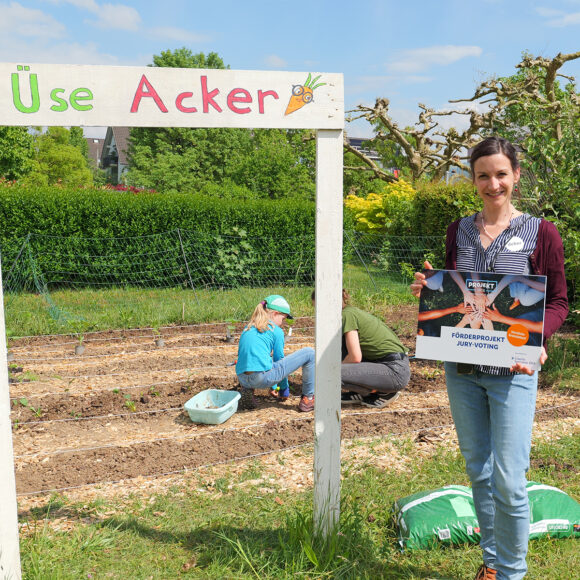 Woman stands in a garden holding a sign that reads 'FÖRDERPROJEKT JURY-VOTING' beside children planting in the soil under a wooden frame that says 'Use Acker'.